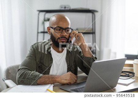 Latin male freelancer calling by cellphone while working online on laptop, having phone conversation at home Latin male freelancer calling by cellphone while working online on laptop, having phone conversation at home 95643518