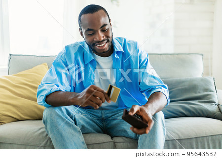 Excited african american man selecting credit card from his card wallet, sitting on sofa in living room interior 95643532