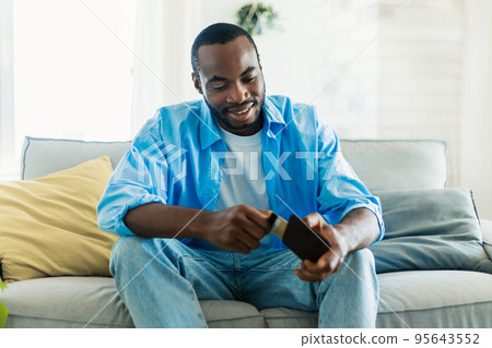 Happy african american man smiling while taking credit card out of wallet, sitting on couch in living room, copy space 95643552