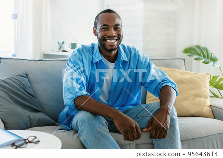 Portrait of handsome black man smiling and looking at camera, sitting on sofa at home, spending time indoors Portrait of handsome black man smiling and looking at camera, sitting on sofa at home, spending time indoors 95643613