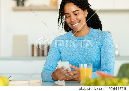 Positive black woman holding smartphone, sitting at kitchen table with glass of fresh juice and vegetables, copy space 95643716