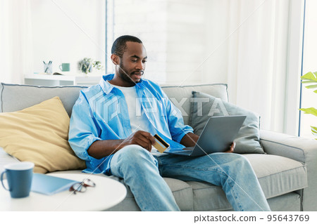 Handsome black man with laptop and credit card sitting on couch, shopping in web store from home, copy space 95643769