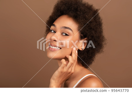 Smiling young mixed race female with curly hair applies cream on face, touches perfect skin with fingers 95643770