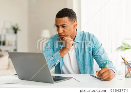 Serious concentrated young black man in jeans looking at laptop, making notes, watching online lesson 95643774