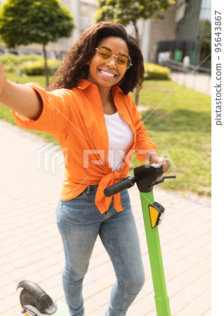 Happy millennial african american lady in casual taking selfie on smartphone on electric scooter in city street 95643867
