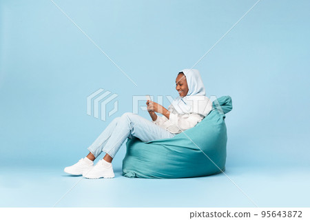 Positive black muslim woman chilling in beanbag chair with smartphone, chatting with friends in social networks 95643872