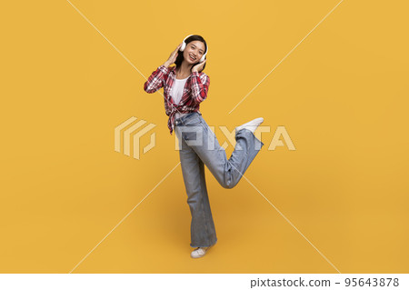 Playful asian lady listening to music in wireless headphones and dancing, having fun over yellow studio background 95643878
