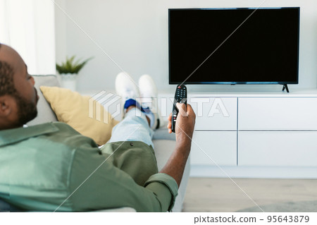Back view of black man watching TV pointing remote control at empty plasma screen, resting on sofa at home, mockup 95643879