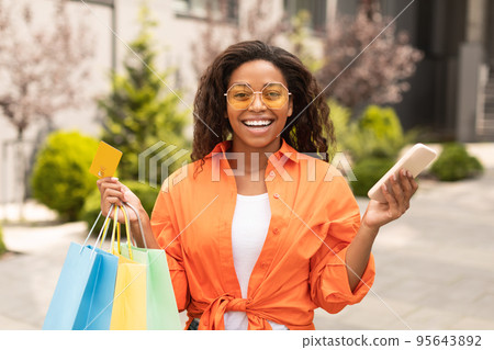 Laughing millennial african american female in casual and glasses with many bags and phone enjoy shopping and walk 95643892