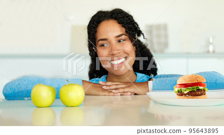 Choice between healthy and unhealthy food. African american woman looking at apple choosing between fruits and junk food Choice between healthy and unhealthy food. African american woman looking at apple choosing between fruits and junk food 95643895