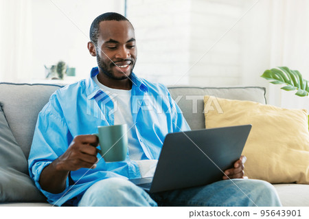 Portrait of smiling black man using laptop, holding cup and drinking morning coffee, sitting on sofa and reading news 95643901