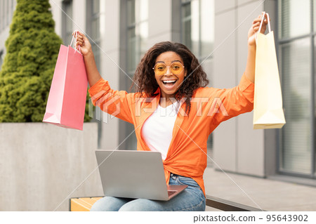 Shocked laughing young african american lady in casual and glasses with laptop picks up many packages, enjoys order 95643902