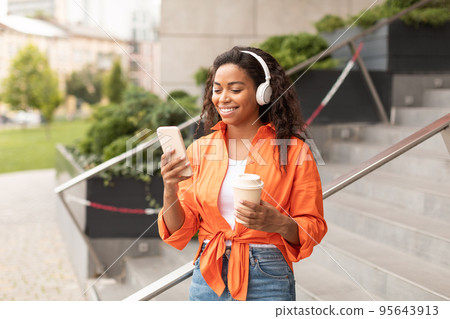 Happy millennial african american lady in casual and wireless headphones chatting on smartphone with cup of coffee Happy millennial african american lady in casual and wireless headphones chatting on smartphone with cup of coffee 95643913