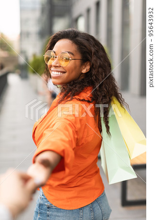 Cheerful pretty millennial african american female with many bags enjoys walk and leads friend by hand Cheerful pretty millennial african american female with many bags enjoys walk and leads friend by hand 95643932