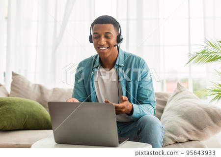 Cheerful young black man in jeans with headphones has video call on laptop, supports client, chats remotely Cheerful young black man in jeans with headphones has video call on laptop, supports client, chats remotely 95643933