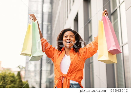 Happy excited pretty millennial african american lady in casual with open mouth with many bags, rising hands up Happy excited pretty millennial african american lady in casual with open mouth with many bags, rising hands up 95643942