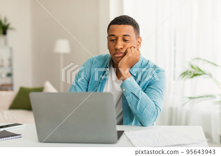 Sad tired bored young black guy in casual looks at laptop in light living room office interior, empty space 95643943