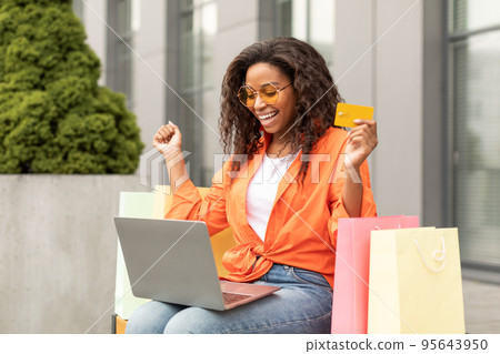 Satisfied millennial african american female in glasses with many bags with purchases, rejoice to win, shows credit card Satisfied millennial african american female in glasses with many bags with purchases, rejoice to win, shows credit card 95643950