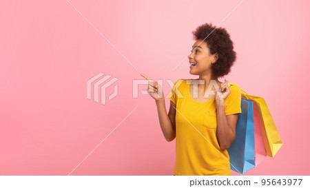 Happy young mixed race female with curly hair with many bags with purchases, points finger at empty space 95643977