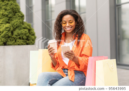 Happy millennial african american lady in casual, glasses with many bags enjoy cup of drink takeaway, typing on phone 95643995