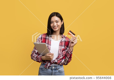 Online shopping concept. Happy chinese lady holding digital tablet and showing credit card, ordering purchases Online shopping concept. Happy chinese lady holding digital tablet and showing credit card, ordering purchases 95644008