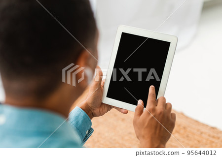 Young black man in casual and wireless headphones chatting on tablet with blank screen in light room Young black man in casual and wireless headphones chatting on tablet with blank screen in light room 95644012