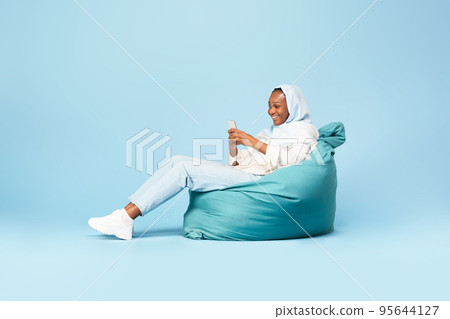 Excited black woman in hijab sitting on beanbag chair and using modern cellphone, using entertaining mobile application Excited black woman in hijab sitting on beanbag chair and using modern cellphone, using entertaining mobile application 95644127