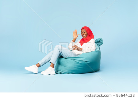 Full length of happy black muslim woman sitting in beanbag chair with smartphone and having video chat with friend 95644128