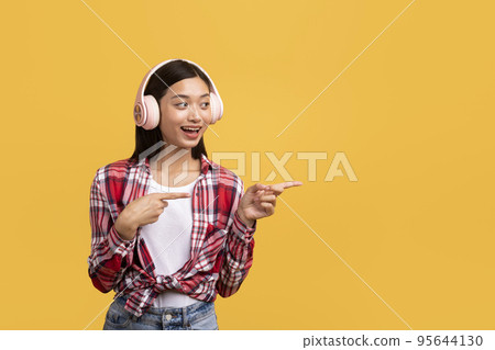 Music lover. Happy asian lady listening to music in headphones and pointing fingers at free space on yellow background Music lover. Happy asian lady listening to music in headphones and pointing fingers at free space on yellow background 95644130
