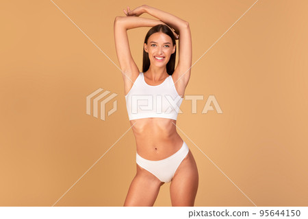 Happy lady in white lingerie posing with hands behind head and smiling at camera over beige background, free space Happy lady in white lingerie posing with hands behind head and smiling at camera over beige background, free space 95644150