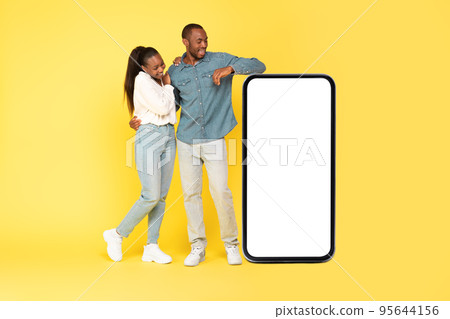 African Couple Looking At Large Phone Embracing On Yellow Background African Couple Looking At Large Phone Embracing On Yellow Background 95644156