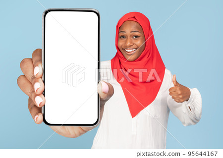 Happy black woman in hijab showing cellphone with white empty screen and gesturing thumb up, mockup 95644167