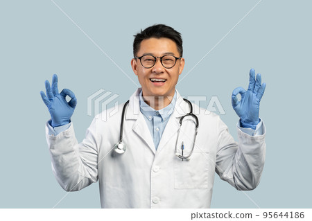 Cheerful middle aged chinese male doctor in white coat, protective gloves, glasses shows ok sign with hands Cheerful middle aged chinese male doctor in white coat, protective gloves, glasses shows ok sign with hands 95644186