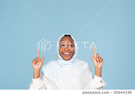 Happy african american muslim woman pointing with both hands and looking up at copy space, showing space for your ad 95644190