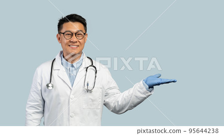 Cheerful middle aged chinese man doctor in white coat, protective gloves, goggles shows hand with empty space Cheerful middle aged chinese man doctor in white coat, protective gloves, goggles shows hand with empty space 95644238