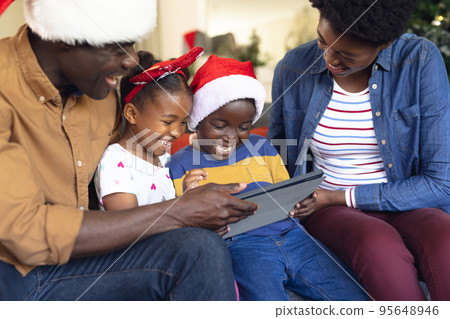 Happy african american family wearing santa hats, using tablet in living room 95648946