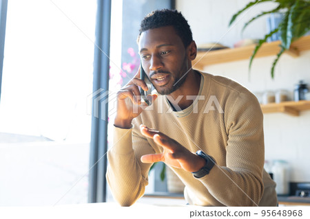 Happy african american man leaning on countertop in kitchen, talking on smartphone 95648968