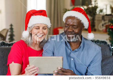 Happy senior diverse couple with santa hats using tablet Happy senior diverse couple with santa hats using tablet 95649658