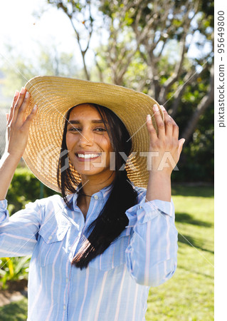 Vertical portrait of happy biracial woman in sunhat standing in sunny garden smiling 95649800