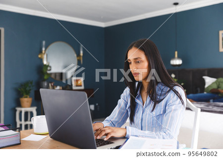 Biracial woman working in bedroom using laptop, with partner in the background 95649802
