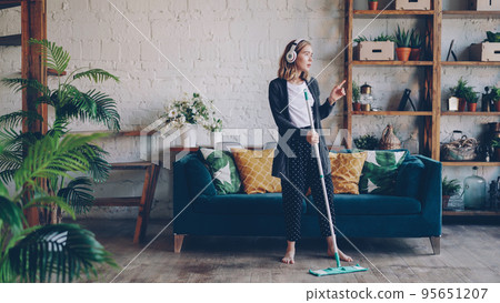 Beautiful young lady is listening to music through wireless headphones and doing housework cleaning the floor with mop. Girl is dancing and singing enjoying cleanup at home. Beautiful young lady is listening to music through wireless headphones and doing housework cleaning the floor with mop. Girl is dancing and singing enjoying cleanup at home. 95651207