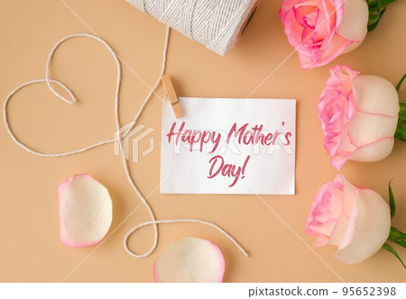 Tender pink roses with spool of white cotton rope in heart shape on beige background. Paper note with text HAPPY MOTHERS DAY. Neutral earth tones. Greeting card 95652398