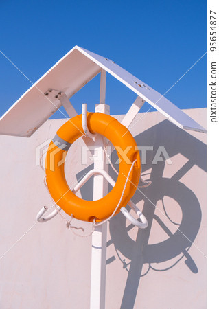 Orange lifebuoy on beach by sea. Safety equipment for rescuing people on seashore. Orange lifebuoy on the pier of blue sky and azure sea Orange lifebuoy on beach by sea. Safety equipment for rescuing people on seashore. Orange lifebuoy on the pier of blue sky and azure sea 95654877
