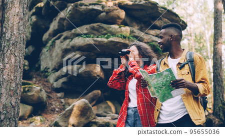 Happy brunette with curly hair is looking through binoculars standing in forest near rocks while her male friend is looking at map, young people are talking. 95655036