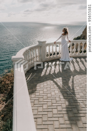 Romantic bride, blond girl in white wedding dress with open shoulders posing on open terrace with backdrop of the sea and rocks. Stylish young woman standing on terrace and looking on ocean at sunset Romantic bride, blond girl in white wedding dress with open shoulders posing on open terrace with backdrop of the sea and rocks. Stylish young woman standing on terrace and looking on ocean at sunset 95655881