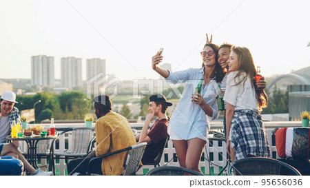 Playful young women are taking selfie with smartphone making funny faces showing tongue and rabbit ears having fun. Beautiful girls are wearing trendy clothes and holding bottles. 95656036