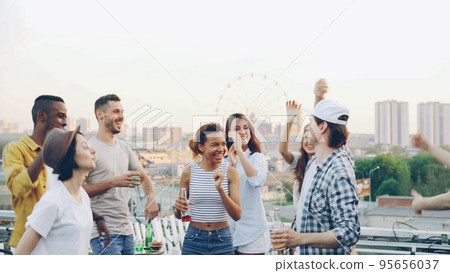 Happy friends are dancing at rooftop party while professional DJ is working with mixing console wearing wireless headphones enjoying rhythm. Beautiful modern city is in background. 95656037