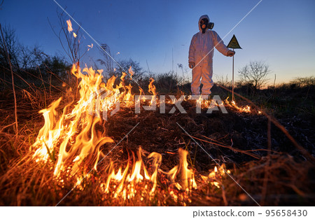 Firefighter ecologist working in field with wildfire at night. Man in suit and gas mask near burning grass with smoke, holding warning sign with skull and crossbones. Natural disaster concept. Firefighter ecologist working in field with wildfire at night. Man in suit and gas mask near burning grass with smoke, holding warning sign with skull and crossbones. Natural disaster concept. 95658430