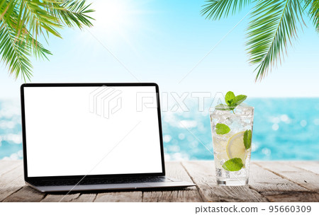 Laptop and cocktail on wooden table in front of sunny sea 95660309