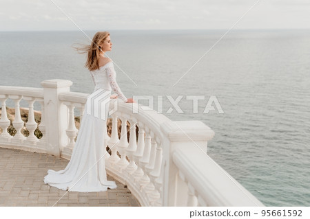 Romantic bride, blond girl in white wedding dress with open shoulders posing on open terrace with backdrop of the sea and rocks. Stylish young woman standing on terrace and looking on ocean at sunset Romantic bride, blond girl in white wedding dress with open shoulders posing on open terrace with backdrop of the sea and rocks. Stylish young woman standing on terrace and looking on ocean at sunset 95661592